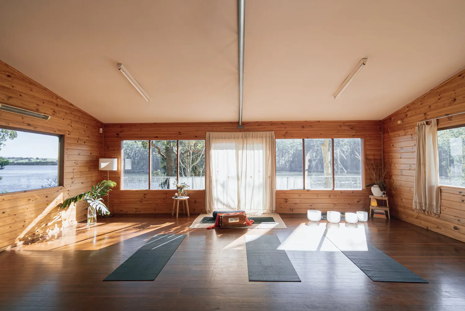 Brown bright wooden room with large windows letting the sunlight in. 3 yoga mats on the floor facing the windows. Looking out the window you can see lots of green trees with water and the sun reflecting off the water as the water sparkles. Theres a box by the window in the middle of the room. A plant on the very left and very right corner of the room. Sound bath bowls on the floor on the right side of the room